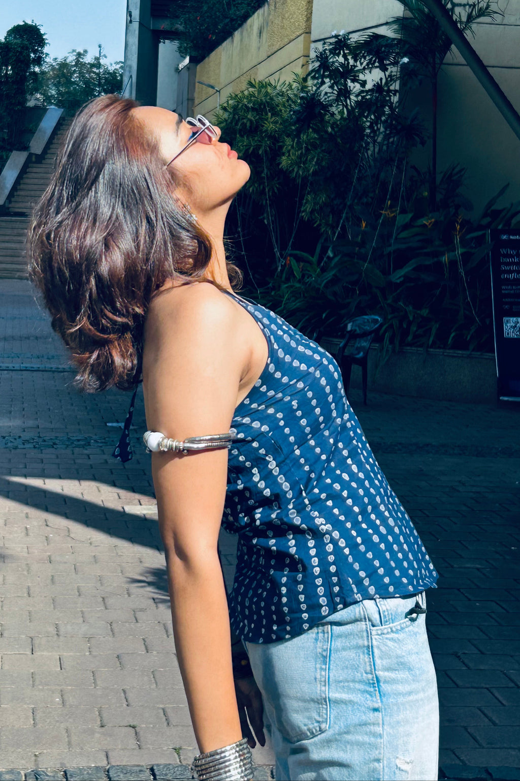 Woman in a blue patterned top and jeans standing outdoors on a sunny day.