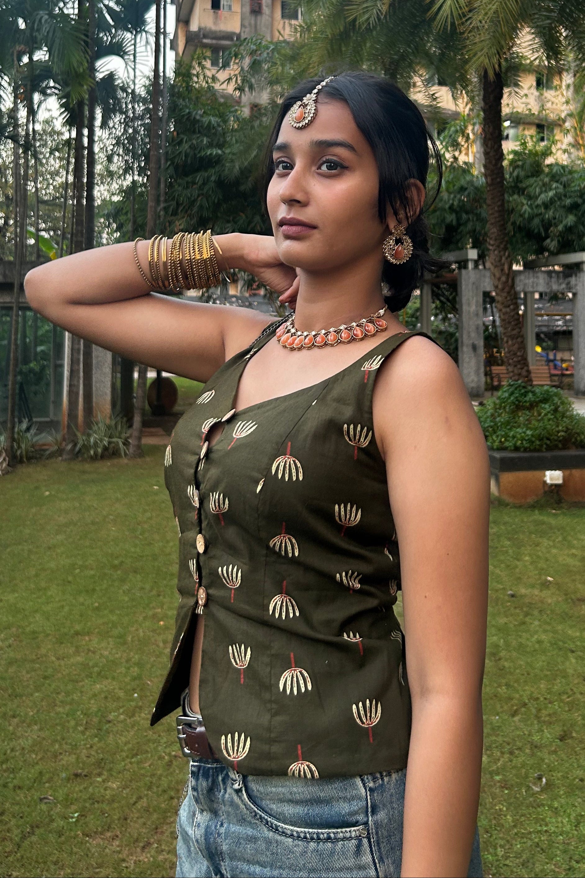 Woman in a green patterned top and blue jeans standing in a garden with palm trees and a building in the background.