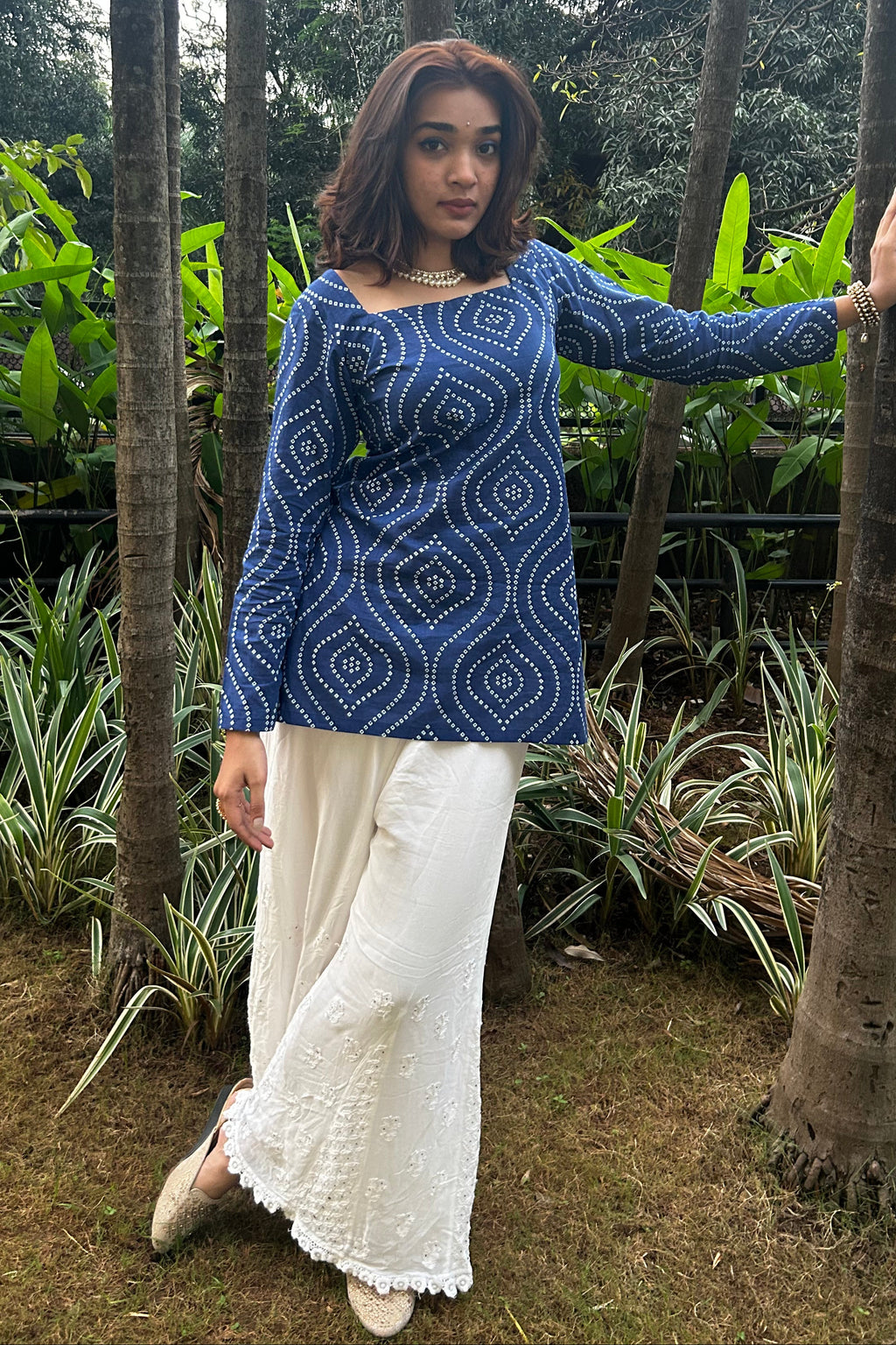 Woman in a blue patterned top and white pants standing among trees and plants.