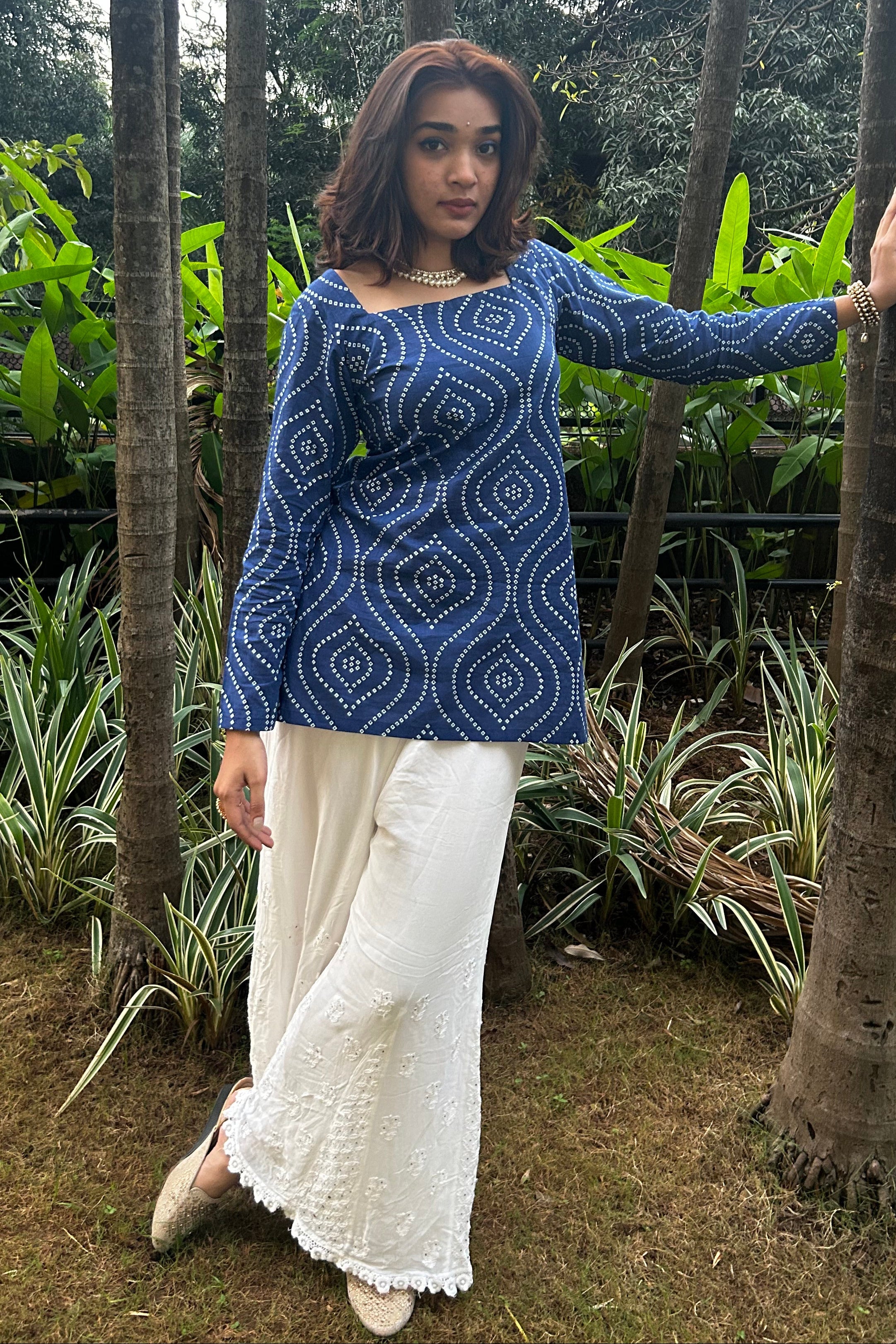 Woman in a blue patterned top and white pants standing among trees and plants.