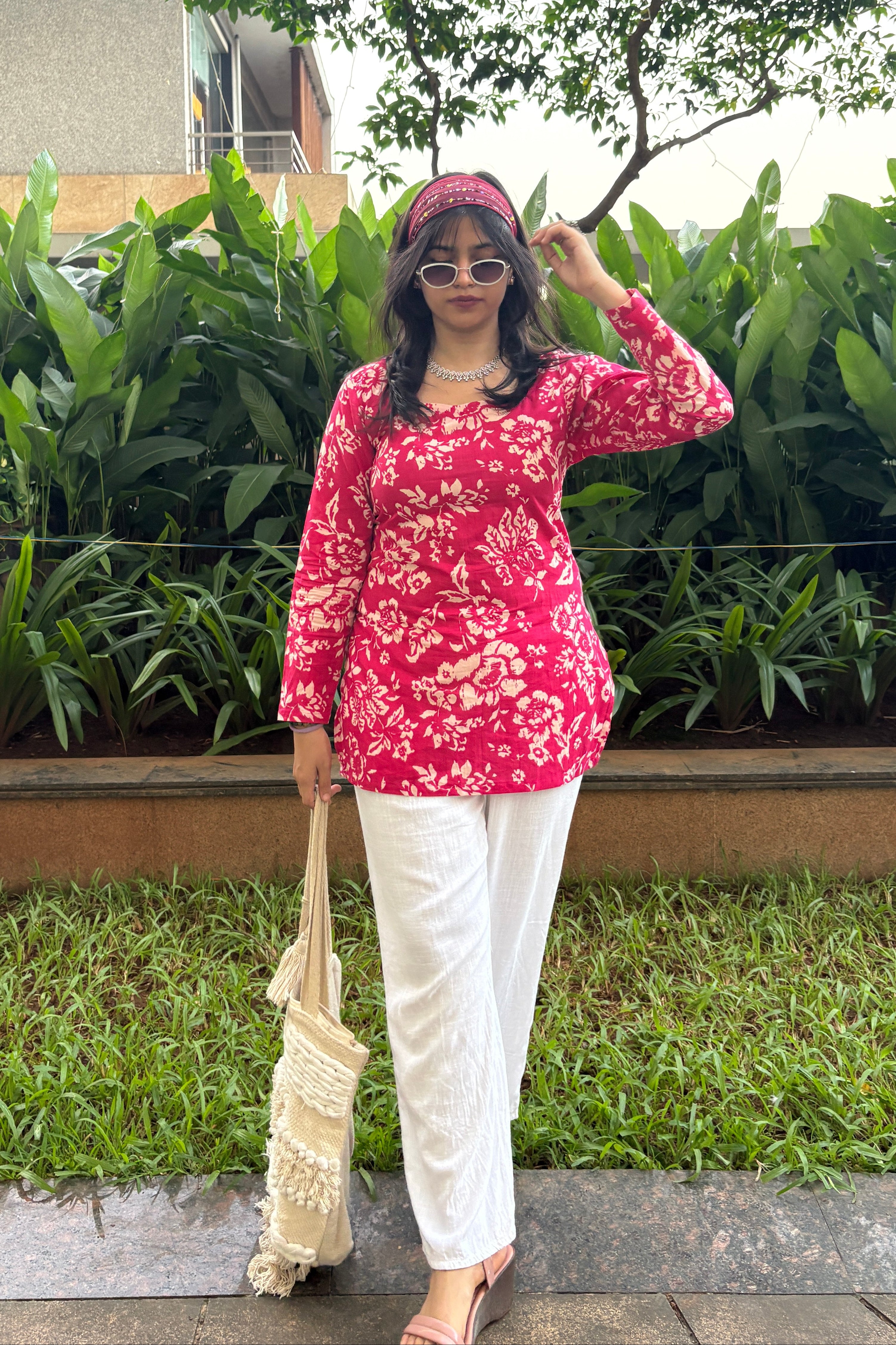 Person in a red floral top and white pants standing outdoors with greenery around