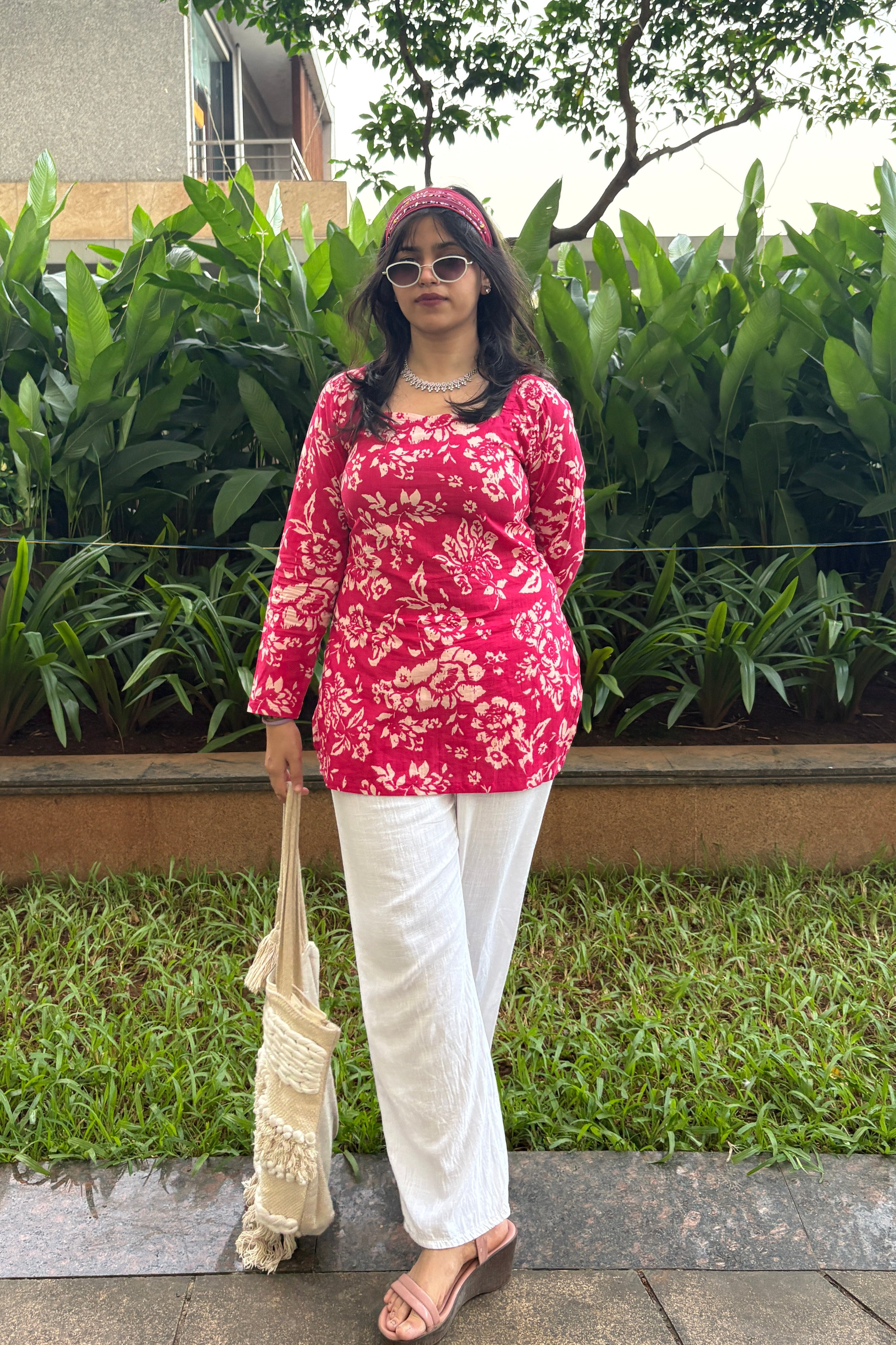 Woman in a red floral top and white pants standing outdoors near greenery