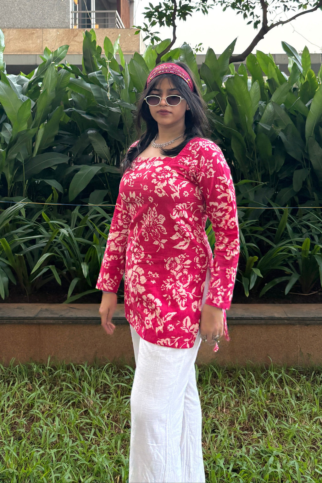 Person wearing a pink floral jacket and white dress standing outdoors with greenery around