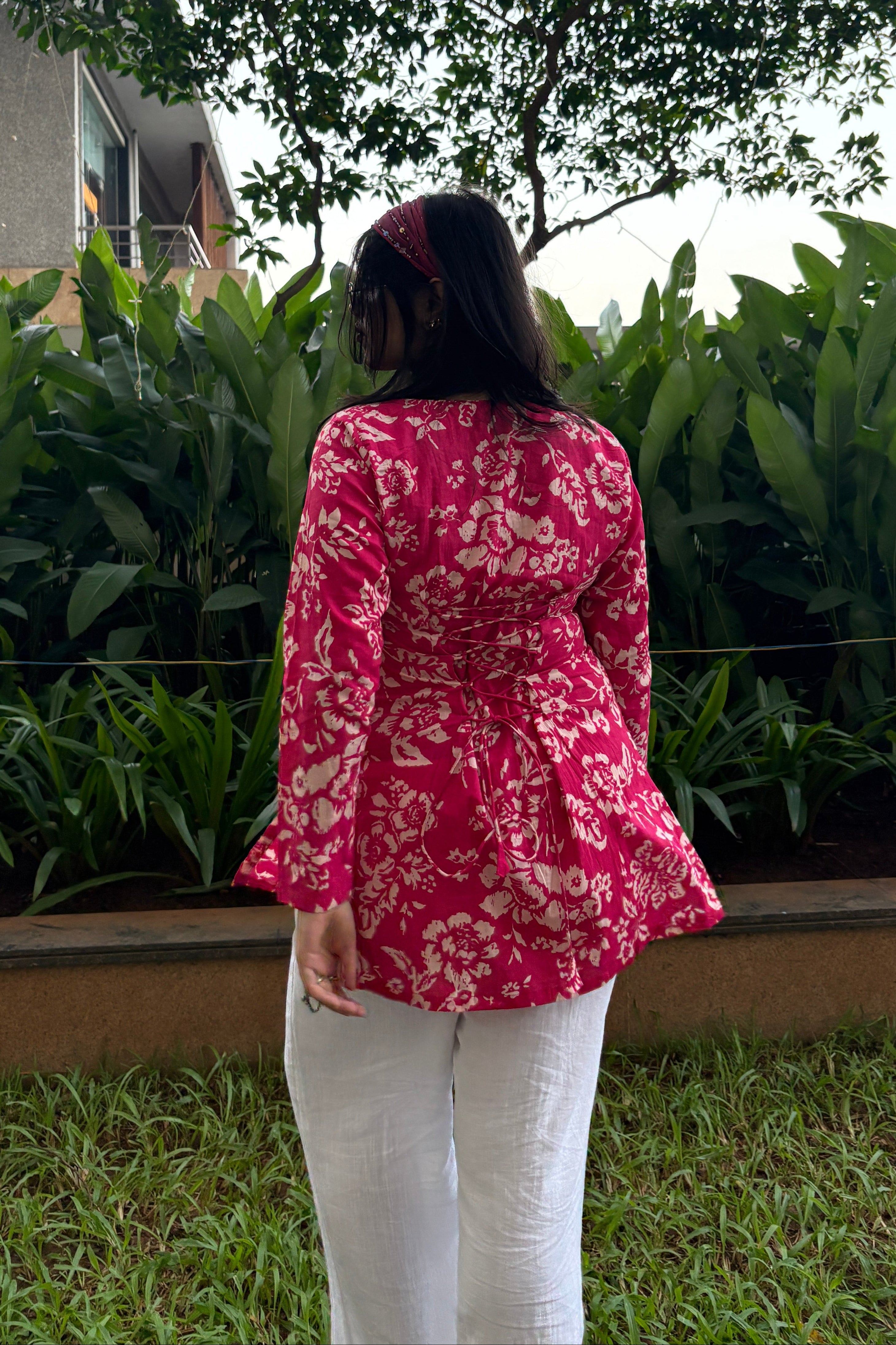 Person wearing a red floral shirt and white pants standing outdoors with greenery around