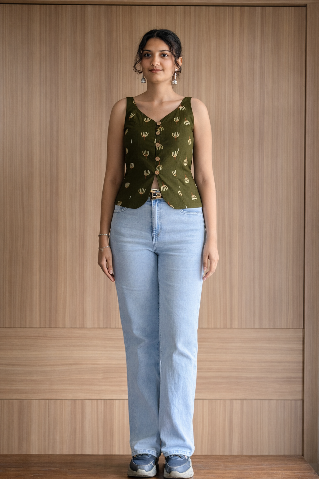 Woman wearing a green patterned top and light blue jeans against a wooden paneled wall.