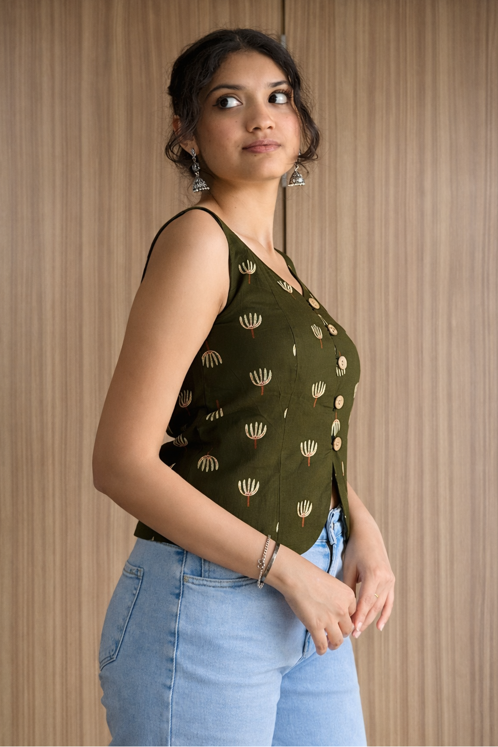 Woman wearing a green sleeveless top with gold patterns and blue jeans against a wooden background