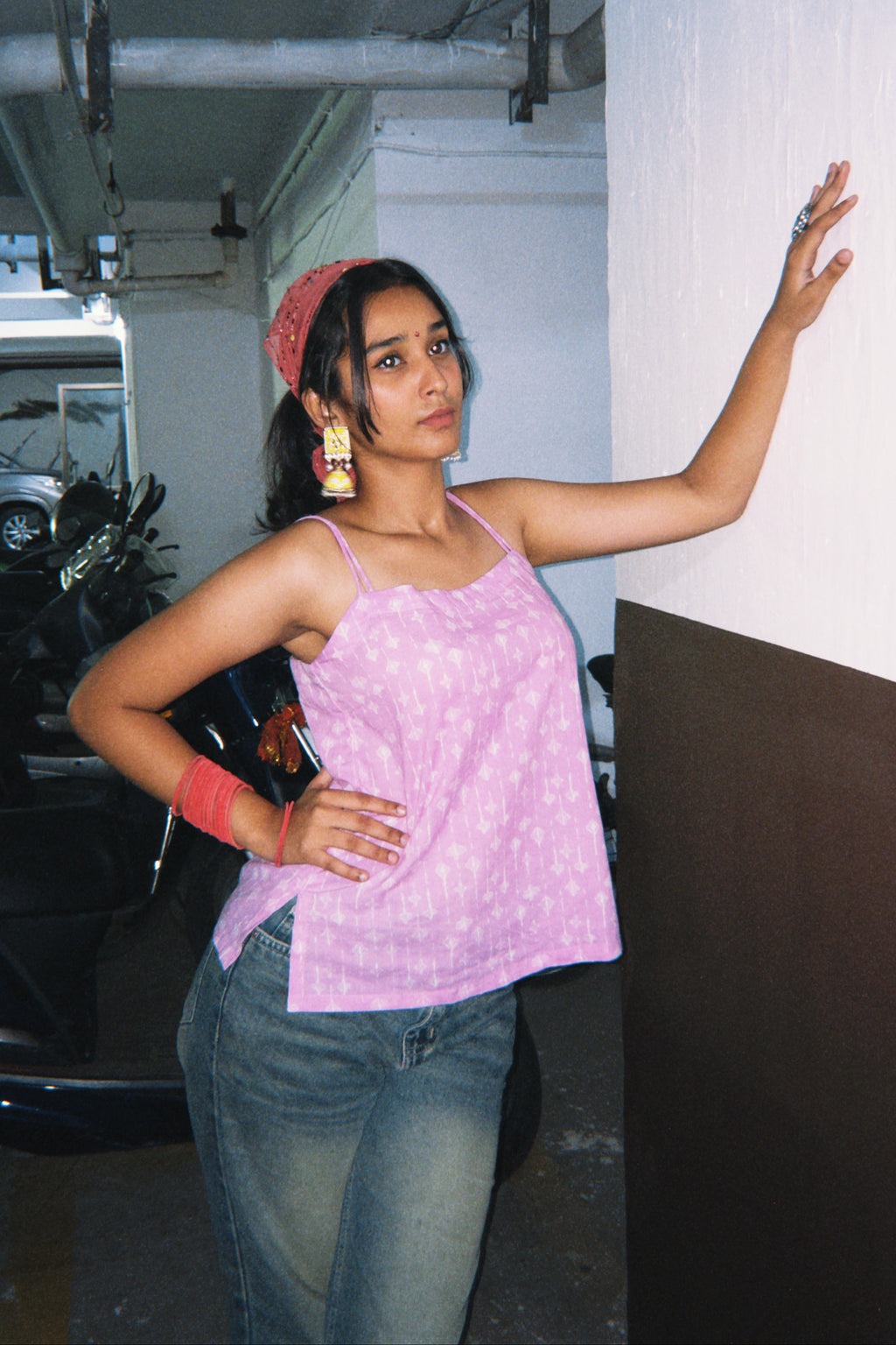 Woman in a pink tank top and jeans standing indoors with a car and wall in the background